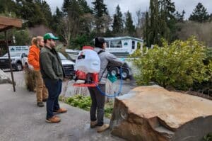 Seattle Tree Care plant health care crew demonstrating growth regulator application equipment at a residential property, with branded company trucks visible in the background.