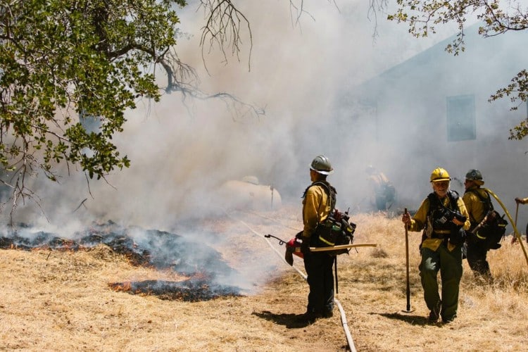 Firefighters controlling a grass fire near a home surrounded by smoke and dry vegetation
